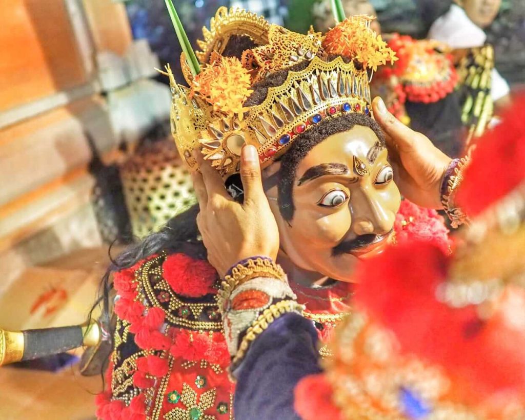 Seorang penari topeng Bali sedang tampil dengan kostum tradisional dan ekspresi dramatis dalam suasana upacara adat.