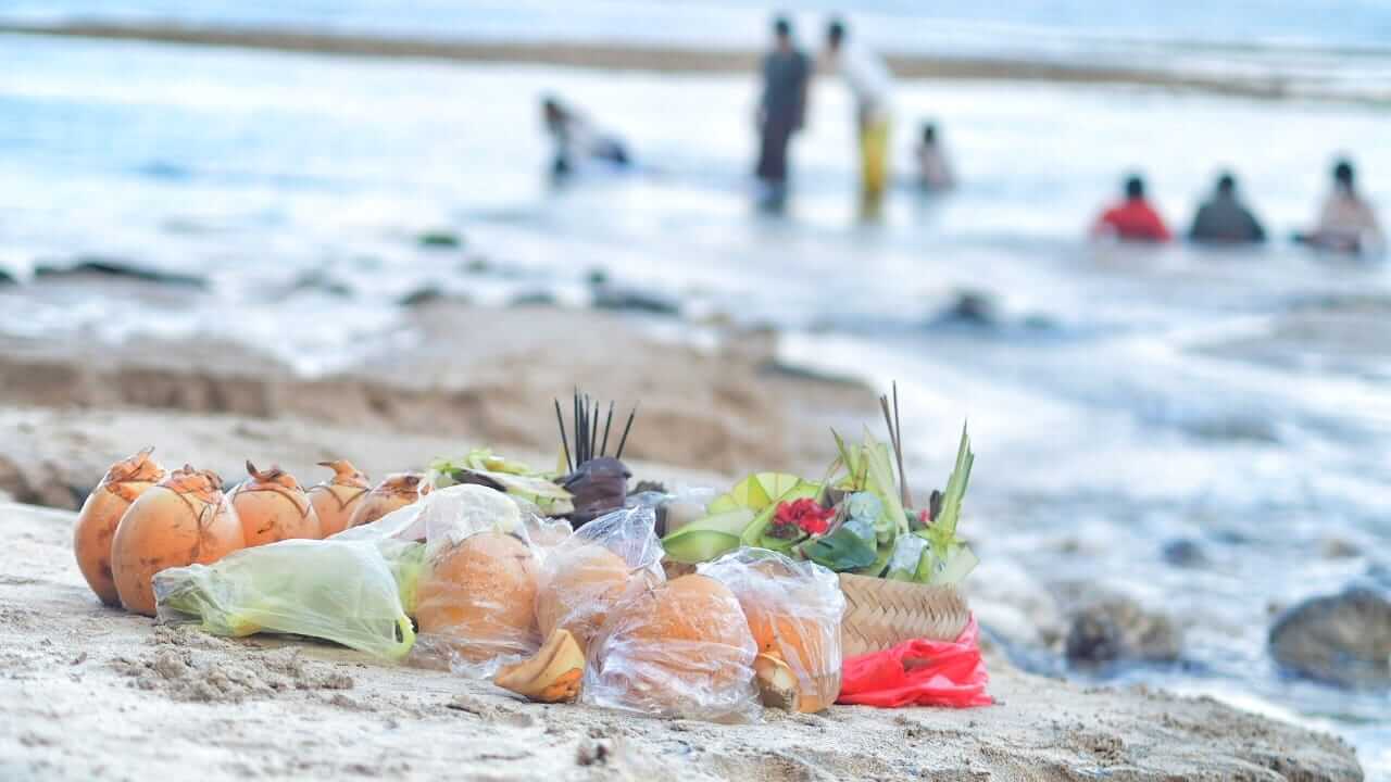Persembahan suci kelapa gading, canang, dan dupa di atas pasir pantai sebagai simbol hubungan manusia dengan semesta.