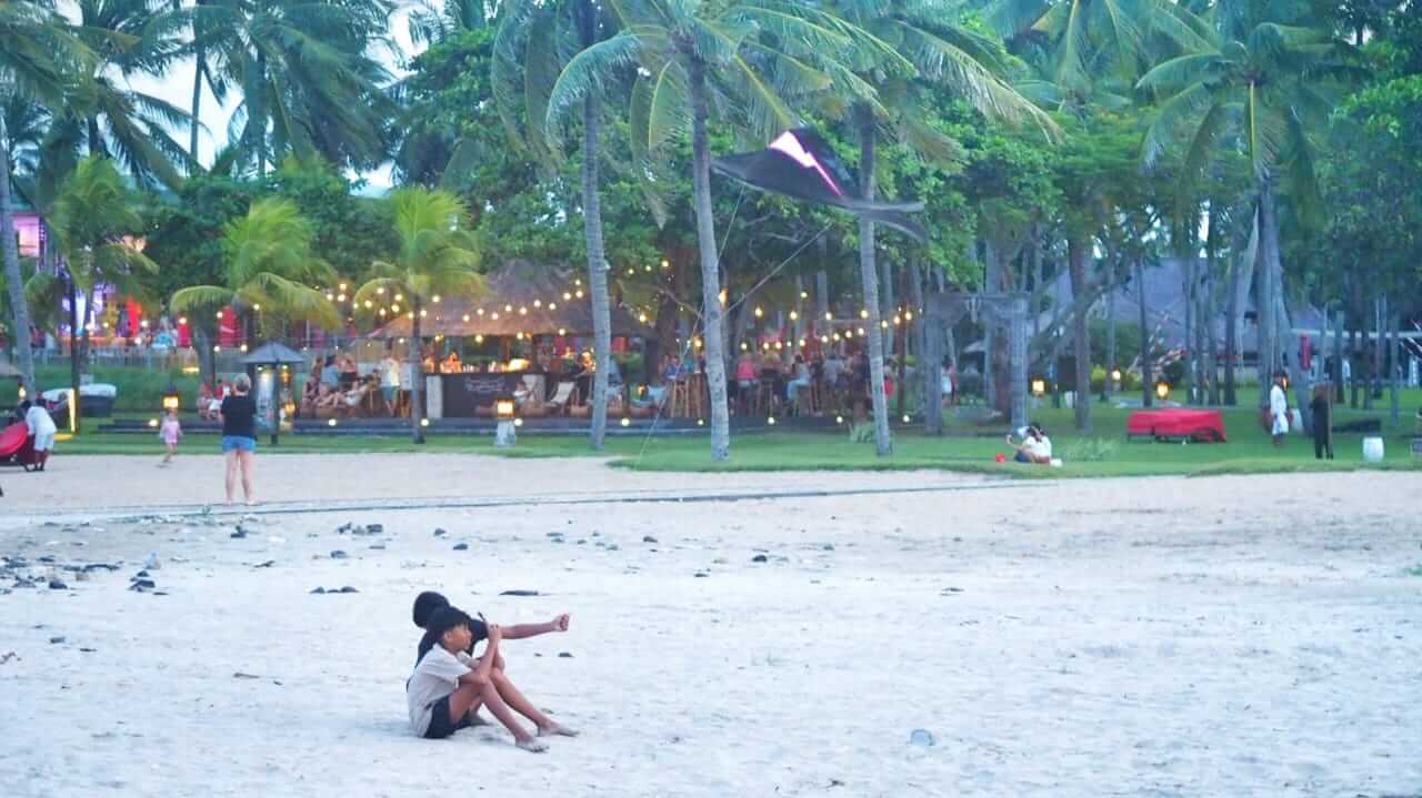 Dua anak laki-laki menerbangkan layang-layang di sisa ruang terbuka tepi pantai, dengan latar belakang gemerlap lampu restoran dan keramaian pengunjung saat senja.