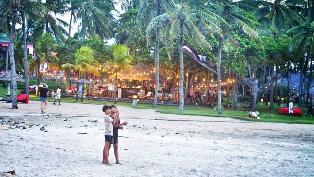 Dua anak laki-laki menerbangkan layangan di tepi pantai senja, menciptakan kontras dengan aktivitas pariwisata yang semakin mendominasi ruang publik di Bali.
