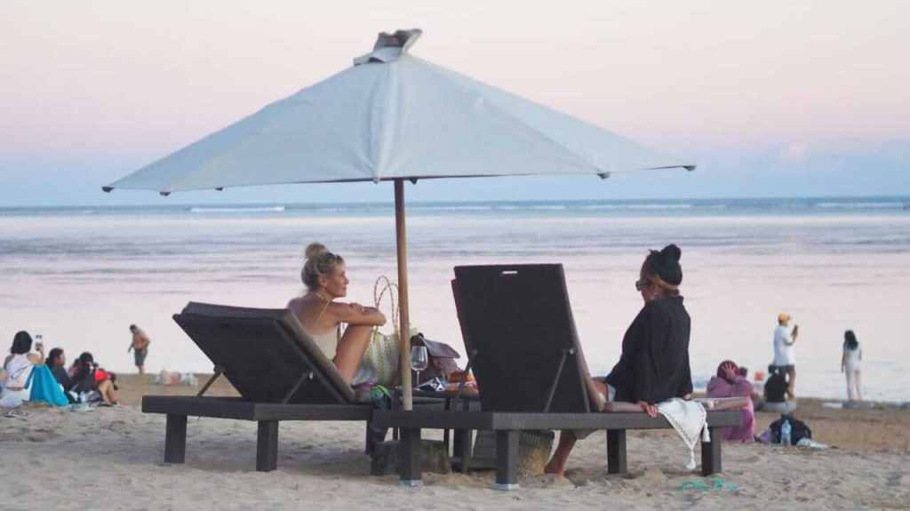 Dua wisatawan bersantai di bawah payung pantai sambil menikmati senja di Bali.