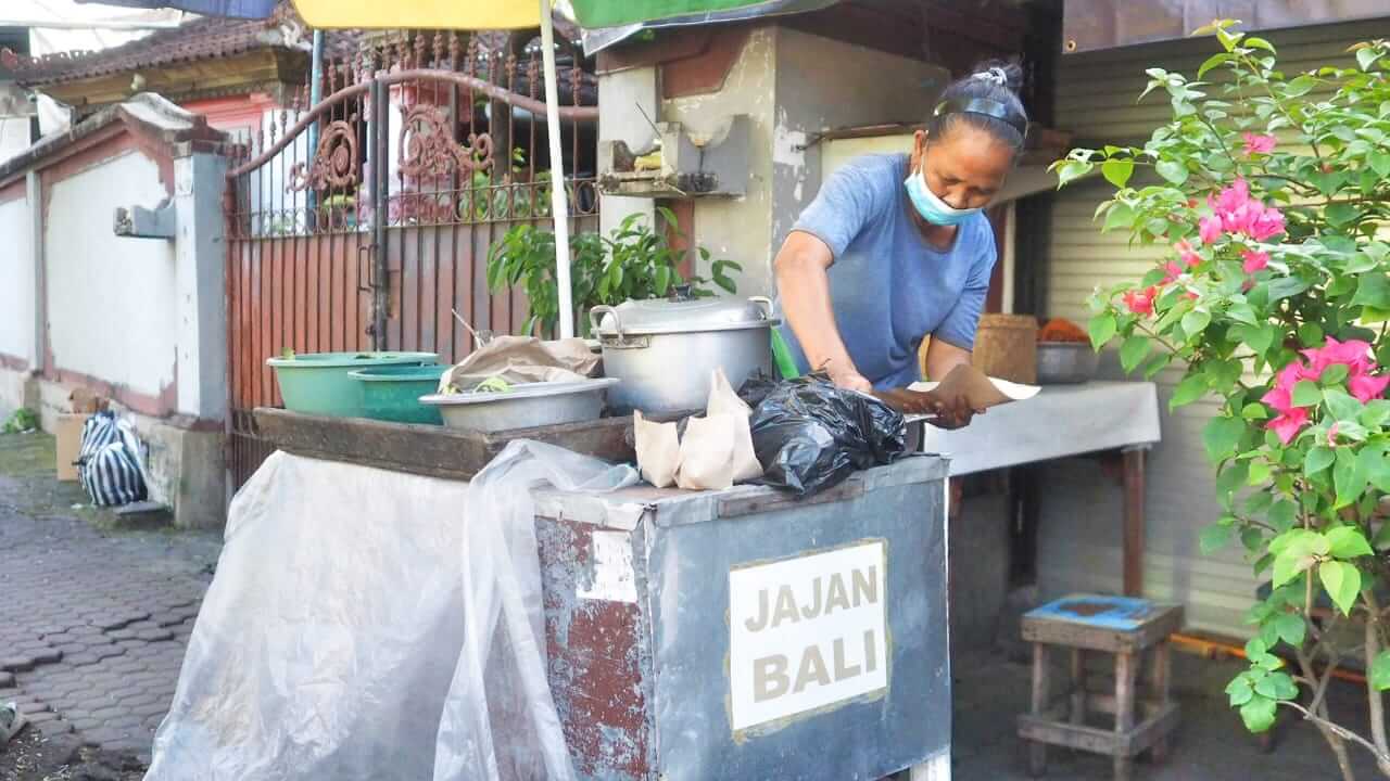 Seorang ibu penjual jajan Bali sedang memasak di dapurnya, melayani pelanggan setia sejak puluhan tahun lalu.