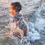 Anak kecil bersama ayahnya bermain ombak di pantai Bali, momen kehangatan dan edukasi di alam terbuka.