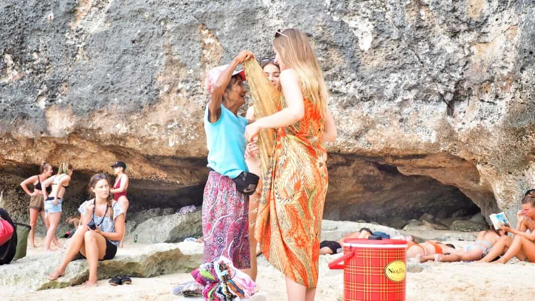 Ibu Bali membantu turis asing memakai kain sambil menjelaskan maknanya dalam bahasa Inggris, cermin interaksi budaya dan pemahaman lintas bahasa.