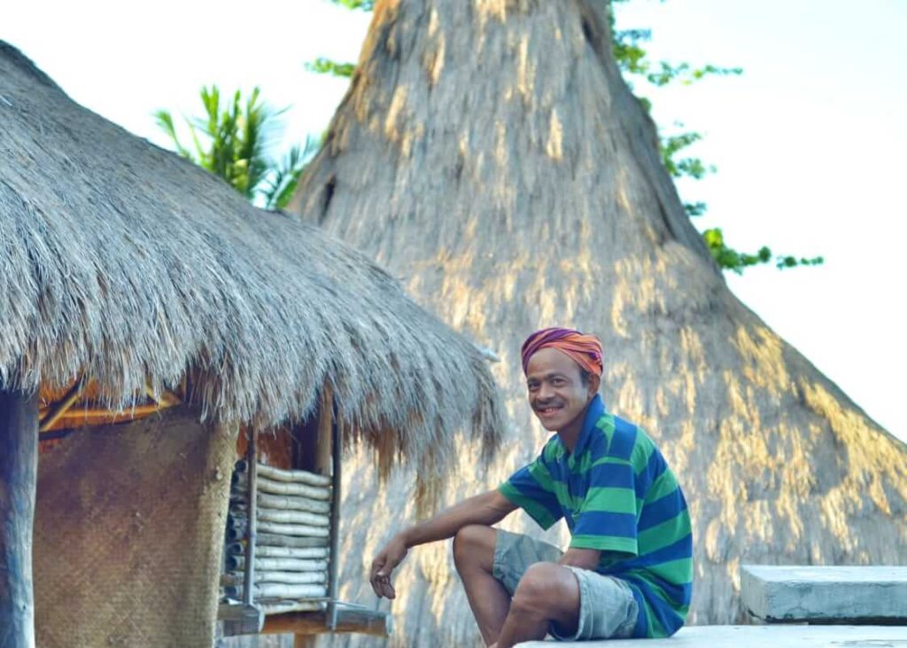 Pria tersenyum di depan rumah adat beratap ilalang di Sumba Barat Daya.