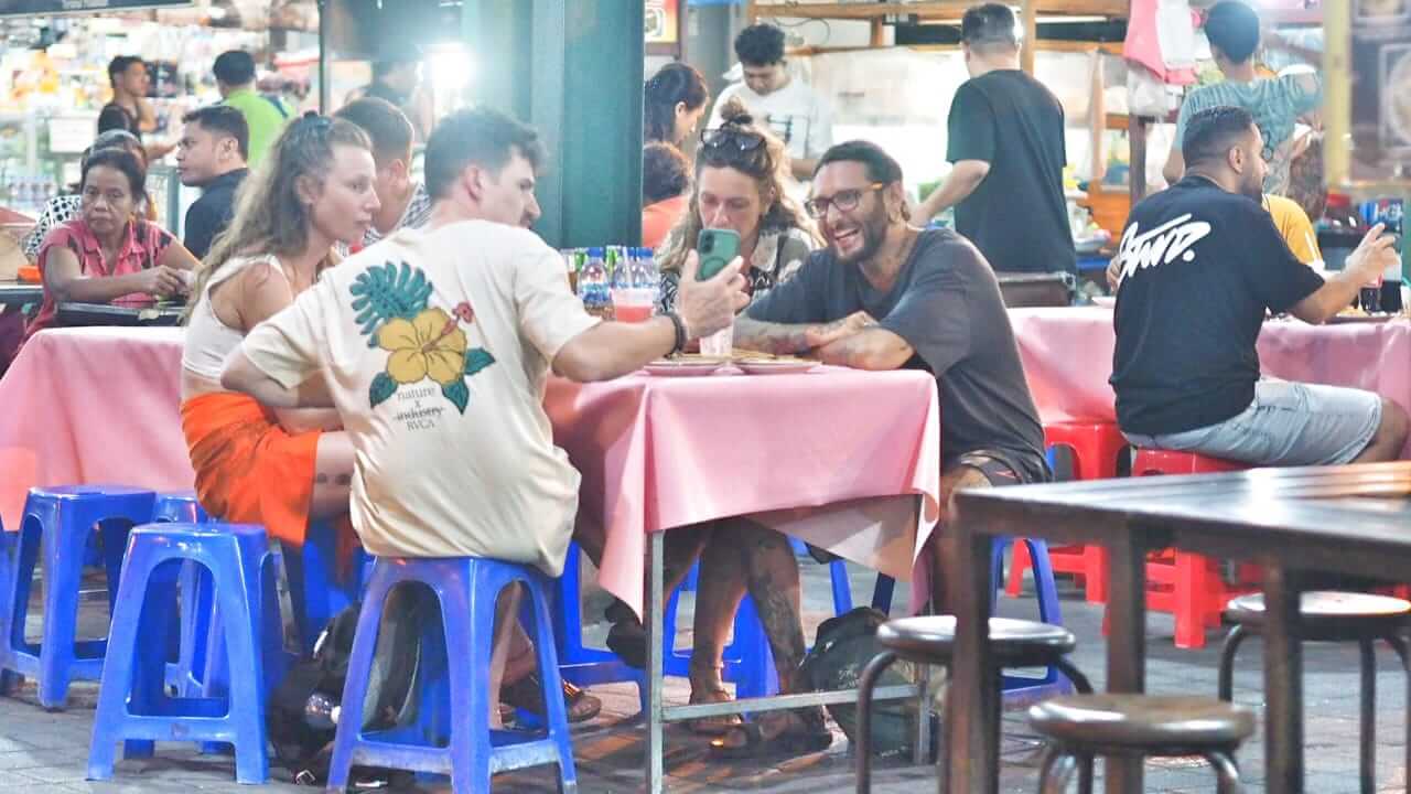 Wisatawan asing menikmati kuliner di pasar tradisional malam Sanur, duduk santai sambil mencicipi beragam hidangan khas Nusantara dalam suasana otentik Bali.