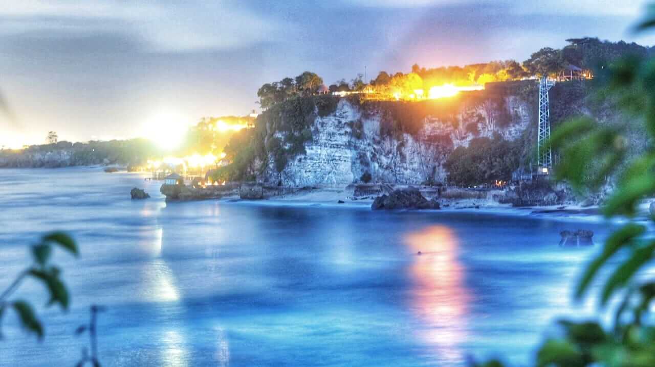 Pemandangan senja di Bukit Jimbaran dengan cahaya lampu dan siluet tebing yang berpadu indah.