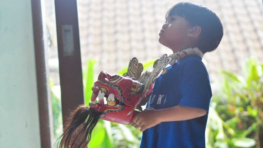 Anak memegang topeng Barong Bali sambil menirukan gerakan dari video YouTube.