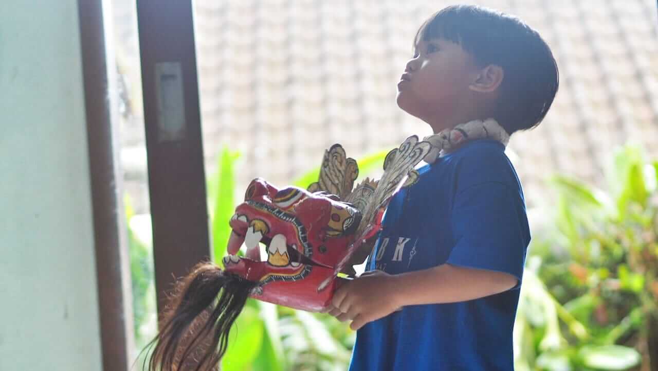 Anak memegang topeng Barong Bali sambil menirukan gerakan dari video YouTube.