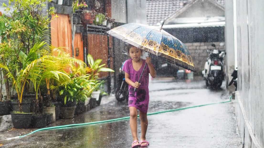 Seorang anak ceria berjalan di tengah hujan sambil membawa payung berwarna.