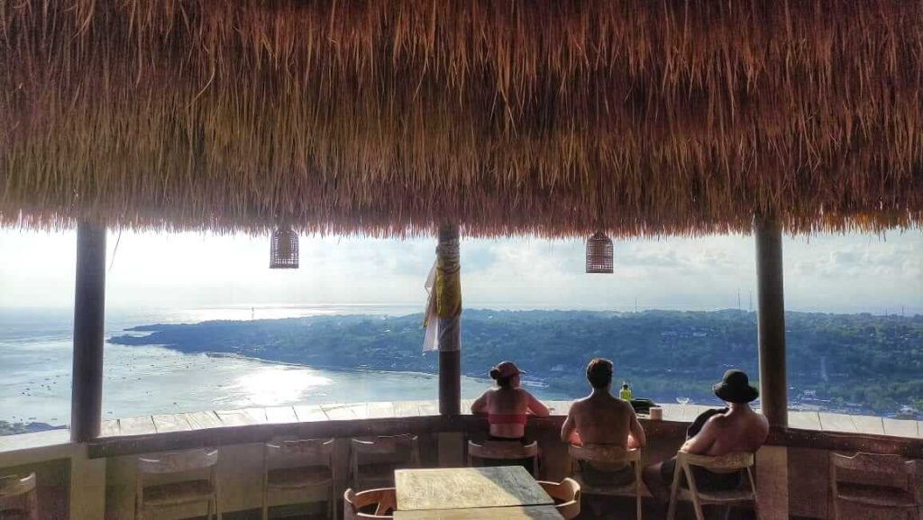 Wisatawan menikmati suasana kafe dengan pemandangan pulau dan pantai dari atas bukit.