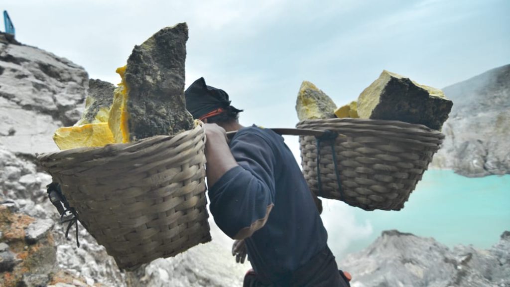 Seorang penambang memikul bambu dengan dua keranjang berisi belerang kuning di kedua ujungnya, menuruni jalur terjal Kawah Ijen.