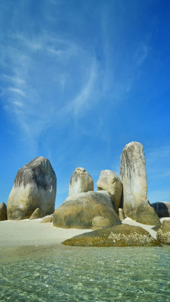 Batu granit besar menjulang di tengah laut dengan hamparan pasir putih dan air jernih.