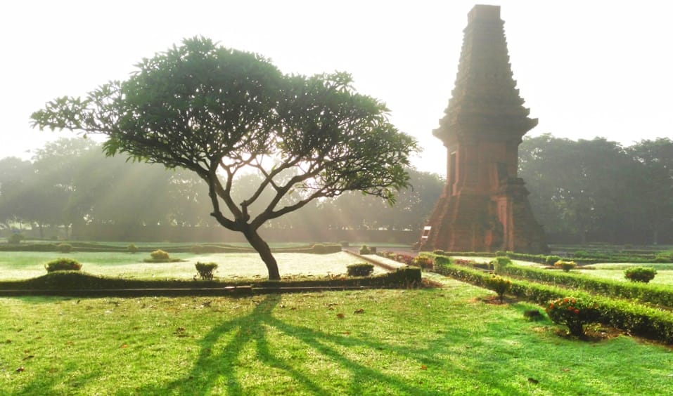 Candi Bajang Ratu di pagi hari dengan cahaya lembut dan suasana tenang, dikelilingi pepohonan hijau.
