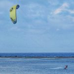 Seorang atlet kite surfing beraksi di laut, memanfaatkan kekuatan angin untuk meluncur di atas ombak.