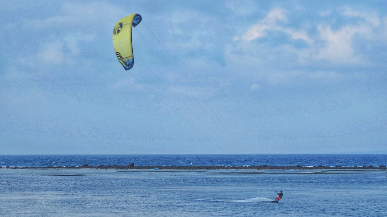 Seorang atlet kite surfing beraksi di laut, memanfaatkan kekuatan angin untuk meluncur di atas ombak.