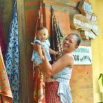 Seorang pria mengenakan busana adat Bali berdiri di area pura, memegang simbol tradisi dengan latar arsitektur khas.