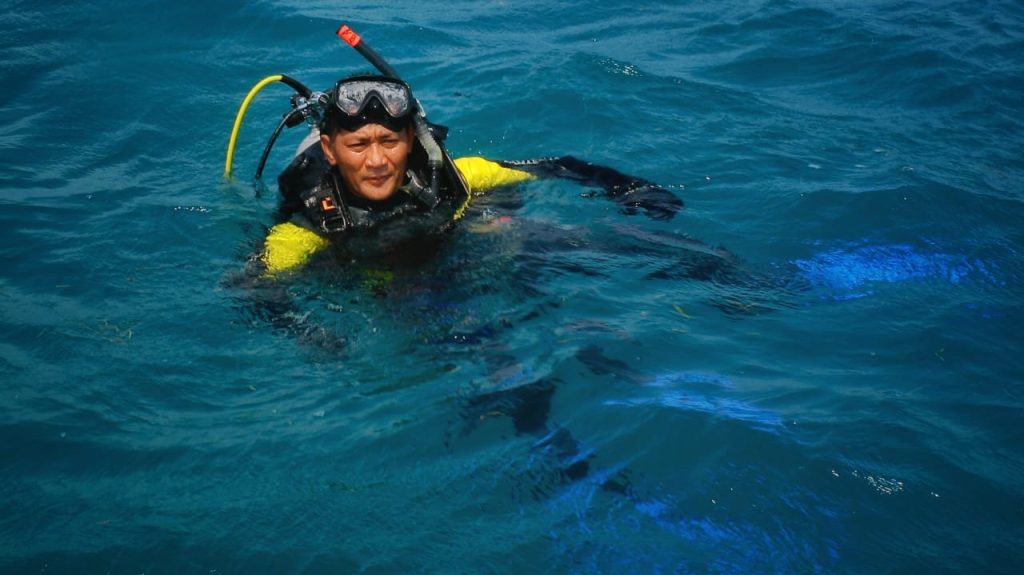 Penyelam berenang di bawah laut dengan perlengkapan lengkap, dikelilingi air jernih dan pemandangan bawah laut.