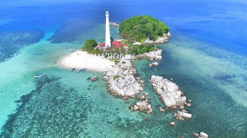 Mercusuar putih menjulang tinggi di Pulau Lengkuas, dikelilingi lanskap alam tropis dan laut biru jernih.