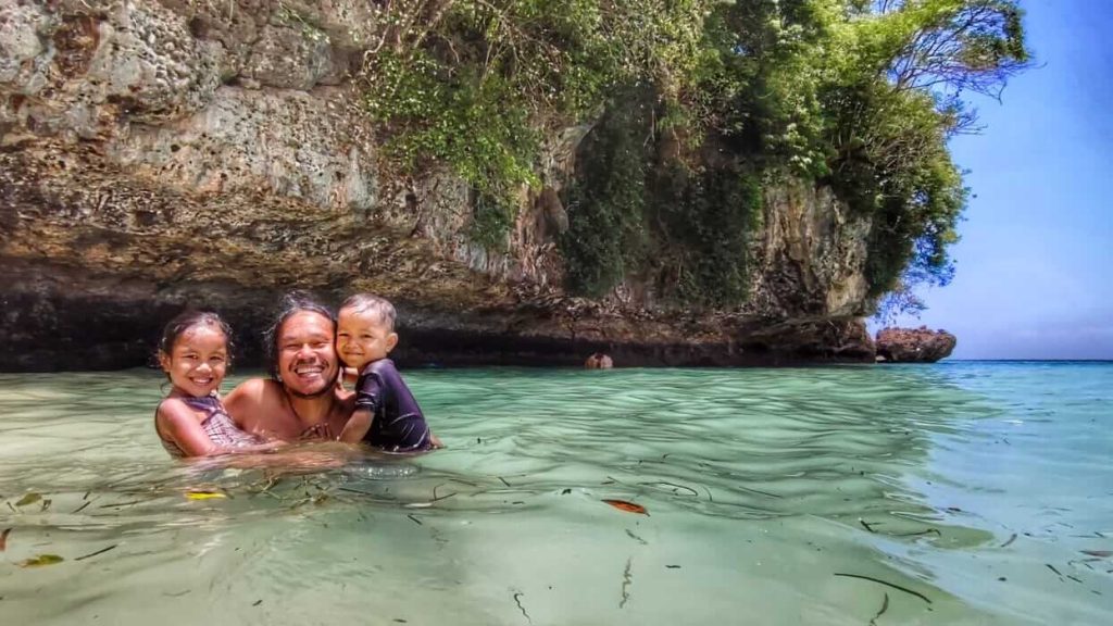 Seorang ayah bermain di tepi pantai bersama dua anaknya, menikmati momen kebersamaan di bawah langit cerah.
