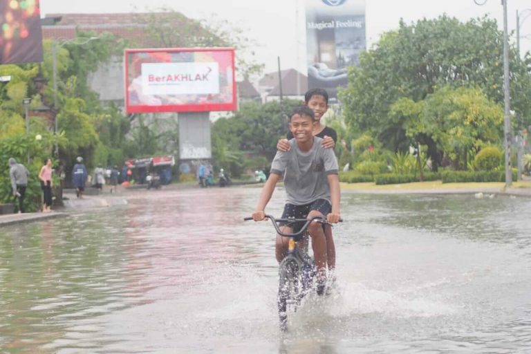 Dua bocah berboncengan sepeda menikmati suasana banjir di jalanan tergenang.