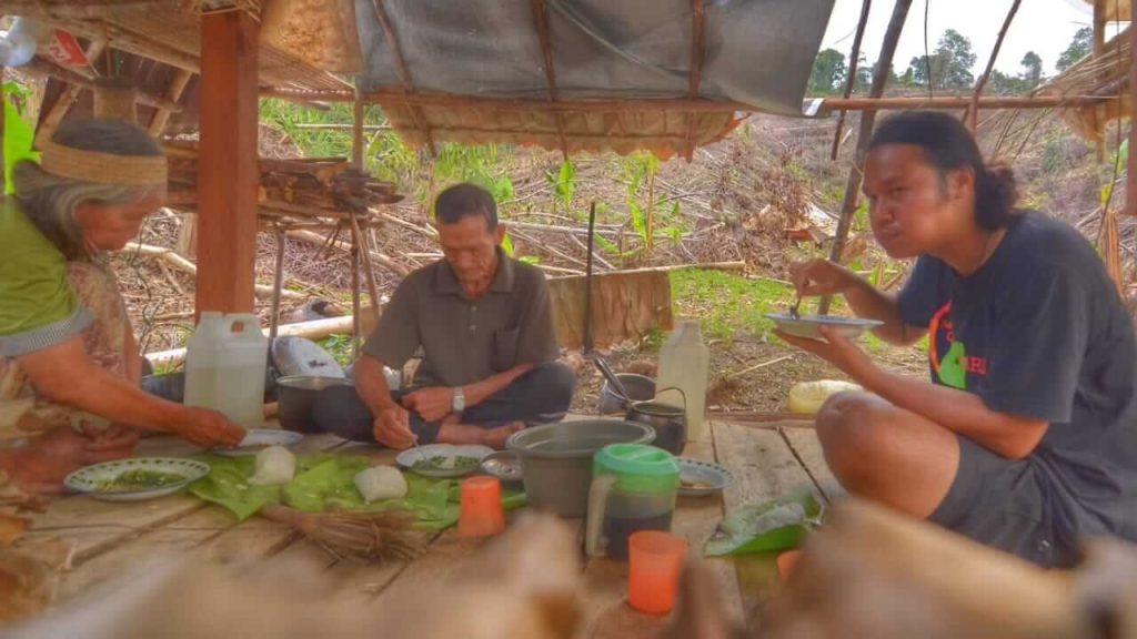 Kakek dan nenek suku Dayak duduk bersama seorang anak muda di pondok kayu, menikmati hidangan tradisional.