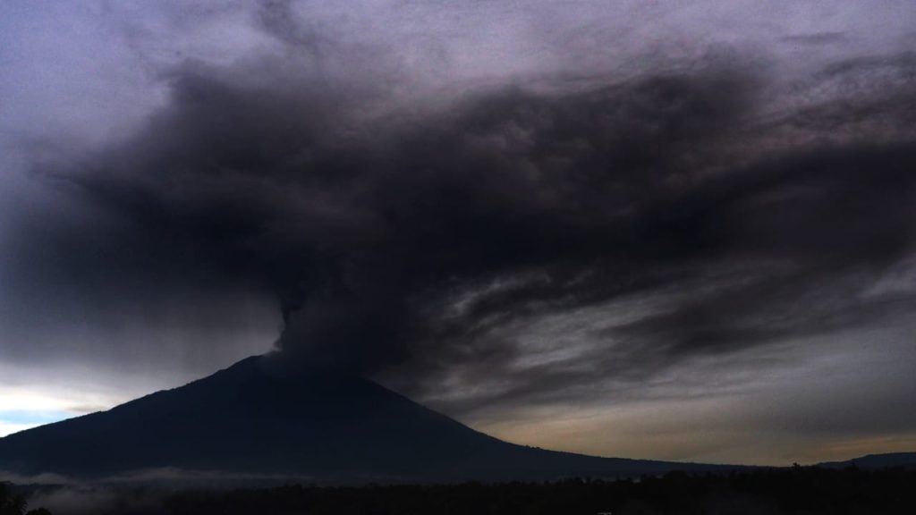 Asap tebal membumbung dari puncak gunung berapi yang sedang meletus di tengah lanskap alam tropis.
