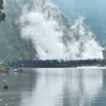 Seorang pemancing menikmati suasana di tepi Danau Segara Anak dengan latar Gunung Rinjani dan api unggun menyala.