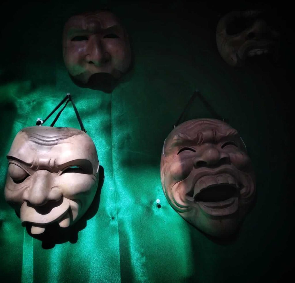 Seorang pria mengenakan topeng dengan ekspresi datar, berdiri di tempat remang.