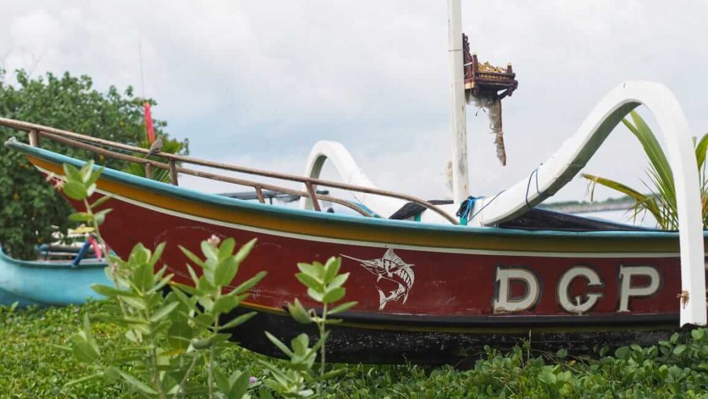 Sebuah jukung tradisional Bali terparkir di tepi pantai, berdiri kontras dengan perkembangan modern di sekitarnya.