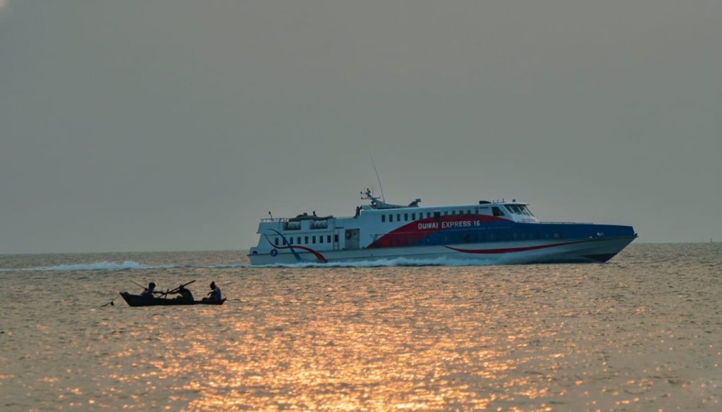 Siluet seorang nelayan tradisional dengan jukung, dihadapkan dengan kapal modern yang melintas di laut yang sama.