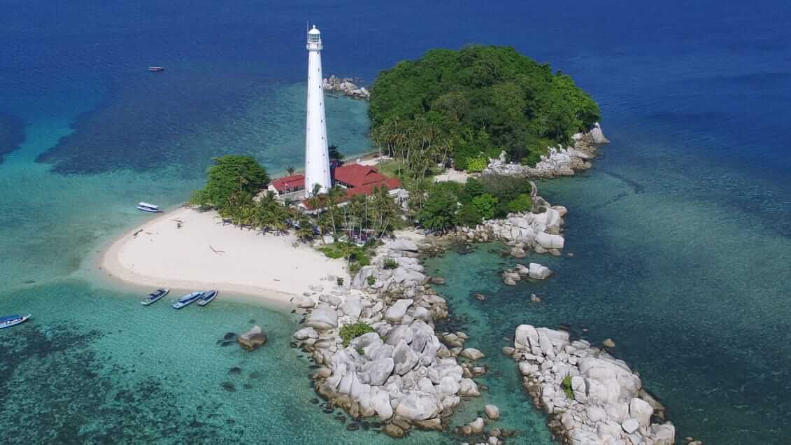 Mercusuar Pulau Lengkuas berdiri megah di tepi laut dengan hamparan batu granit dan air laut biru yang jernih.