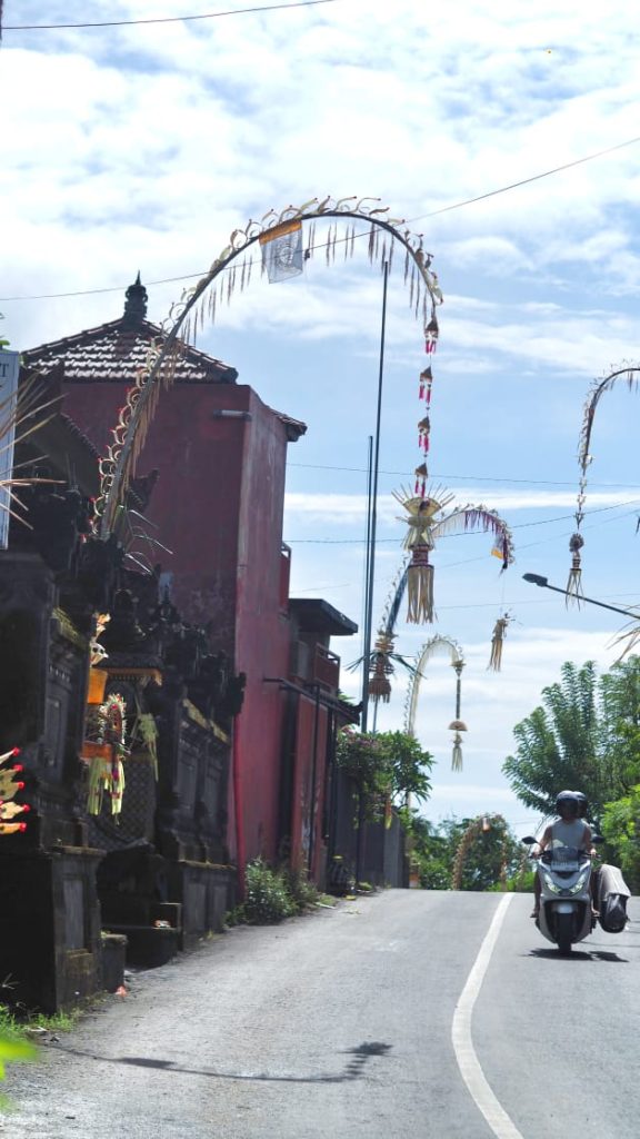 Wisatawan melintas dengan motor sambil membawa papan selancar, melewati deretan penjor yang menghiasi jalan.