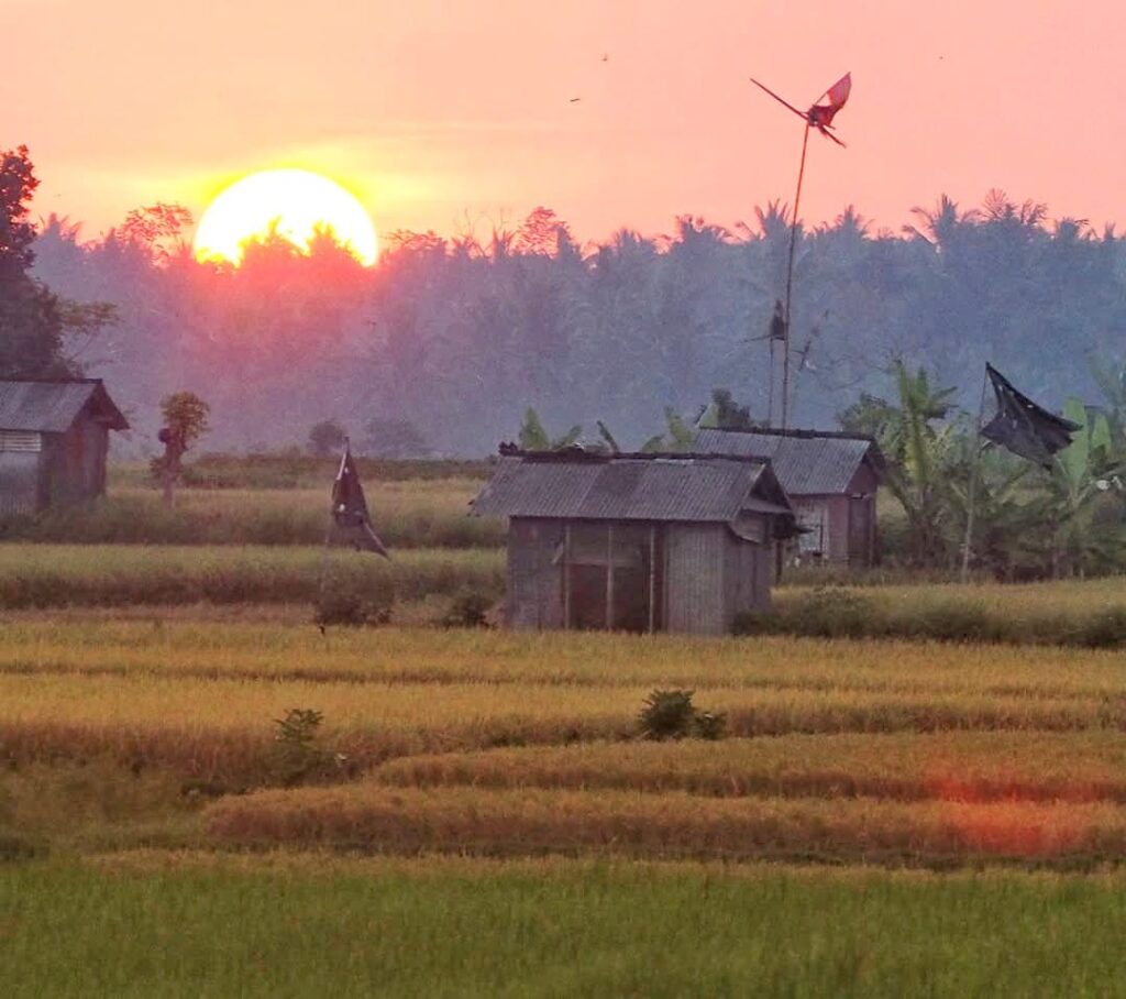 Siluet gubuk kecil dengan latar matahari terbenam, menghadirkan suasana senja yang hening.