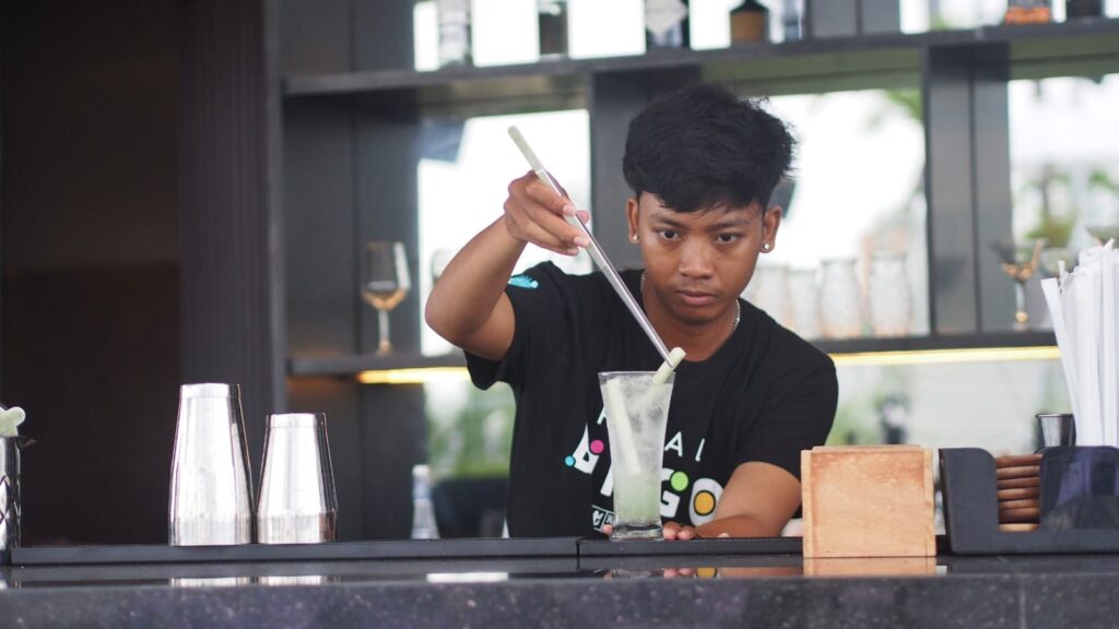 Seorang bartender sedang meracik minuman di bar dengan suasana pencahayaan hangat