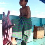 Seorang anak memegang kemudi perahu di atas air dengan suasana sederhana