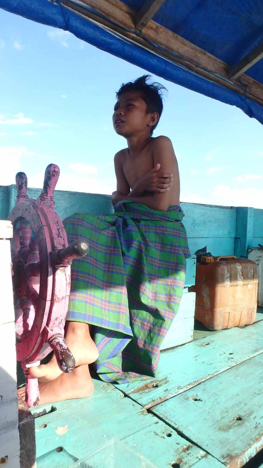 Seorang anak memegang kemudi perahu di atas air dengan suasana sederhana