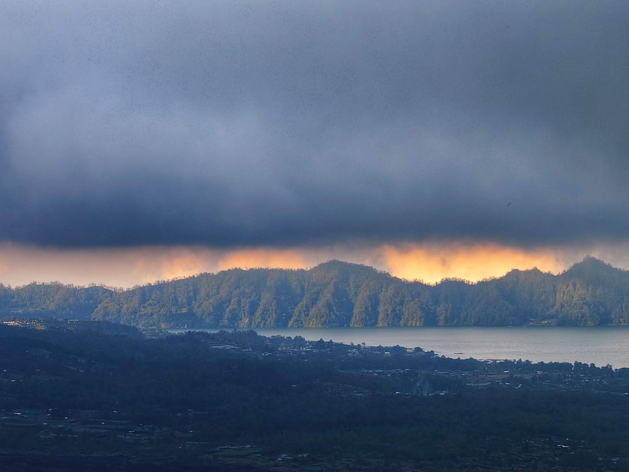 Cahaya jingga muncul di balik awan gelap menciptakan suasana langit yang dramatis
