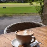Secangkir kopi dengan latar hamparan hijau lapangan golf pada pagi hari.