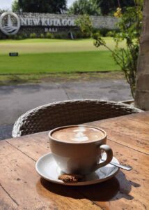 Secangkir kopi dengan latar hamparan hijau lapangan golf pada pagi hari.
